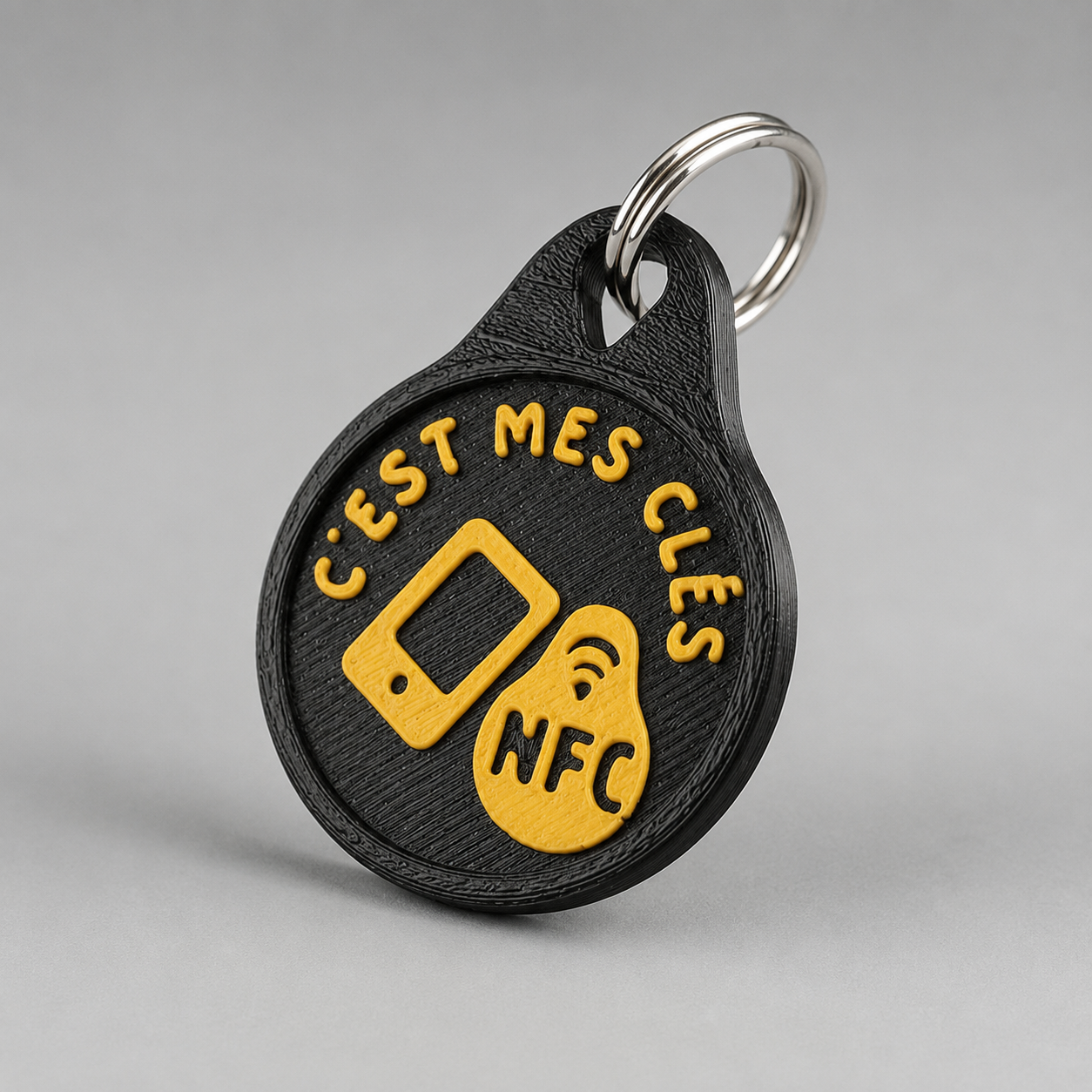 Round black plastic NFC keychain with yellow text reading c'est mes clés and a metal ring on a gray background.
